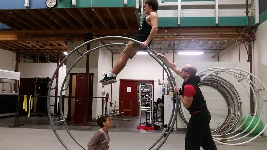 An aerialist practicing on a giant wheel with two helpers assisting